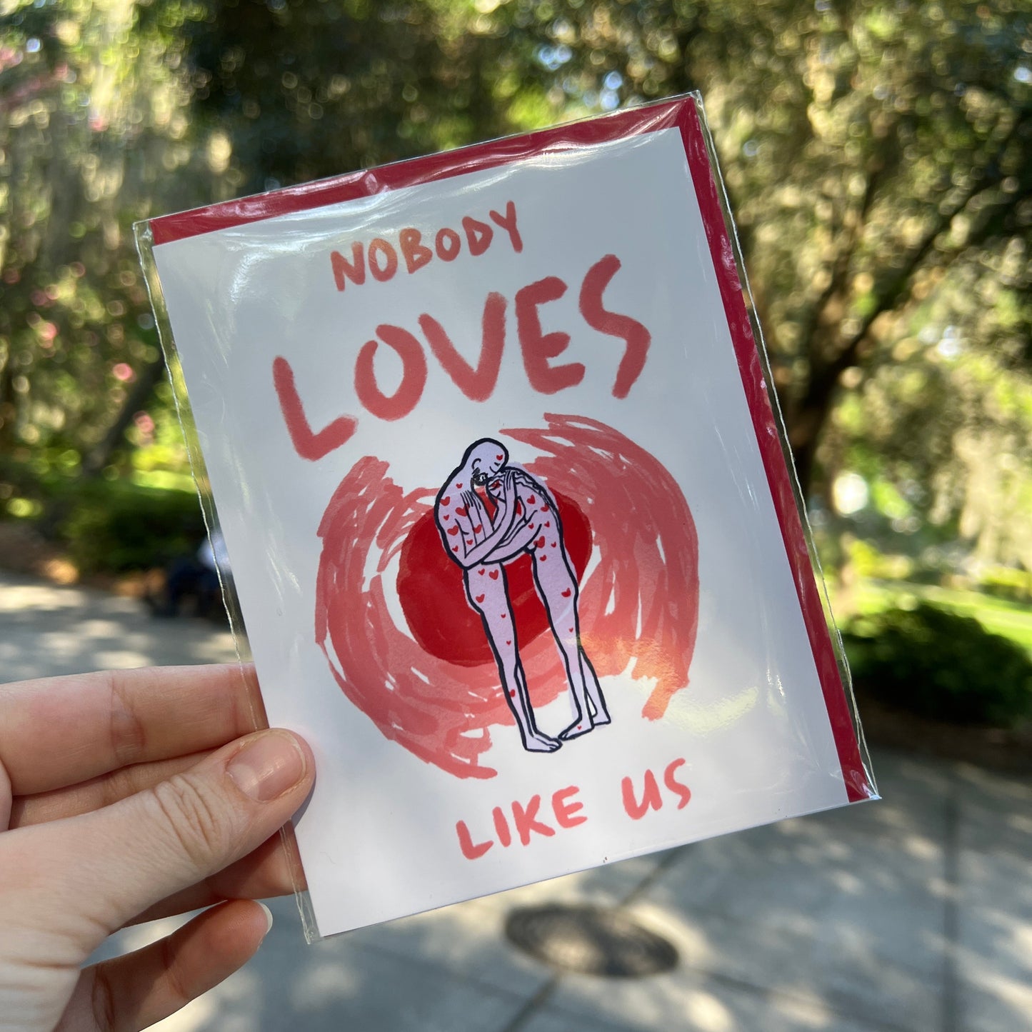 A hand holds a small printed card or sticker in a clear plastic sleeve outdoors. The card has a white background with red border and text reading 'NOBODY LOVES LIKE US' around an illustration of a hugging person inside a red circular mark.