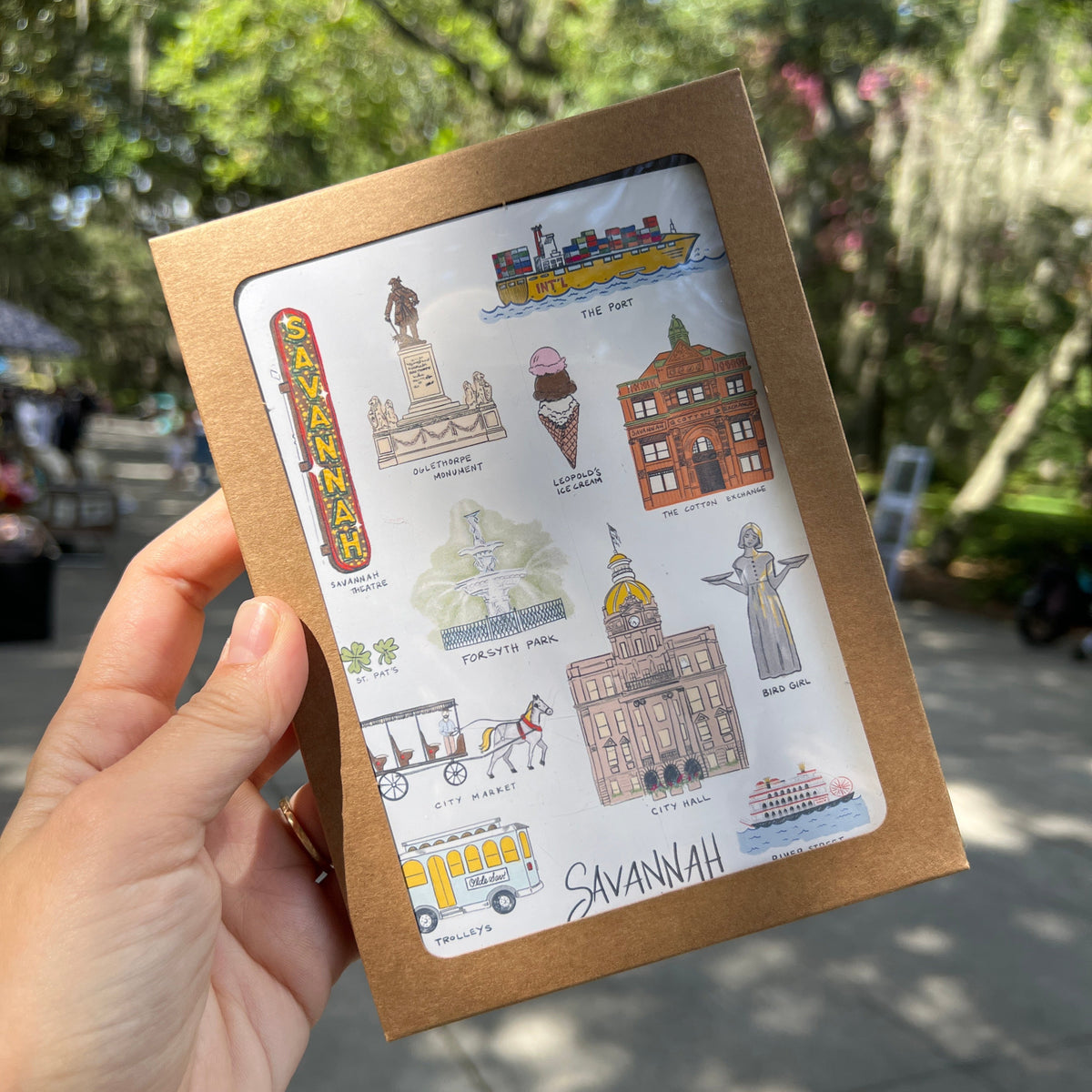 A hand holding a small square kraft paper card or sticker sheet outdoors. The sheet has a white center printed with illustrated landmarks and the word 'Savannah' in script, with a brown cardboard border.