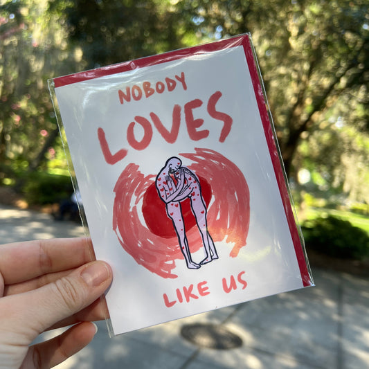 A hand holds a small printed card or sticker in a clear plastic sleeve outdoors. The card has a white background with red border and text reading 'NOBODY LOVES LIKE US' around an illustration of a hugging person inside a red circular mark.