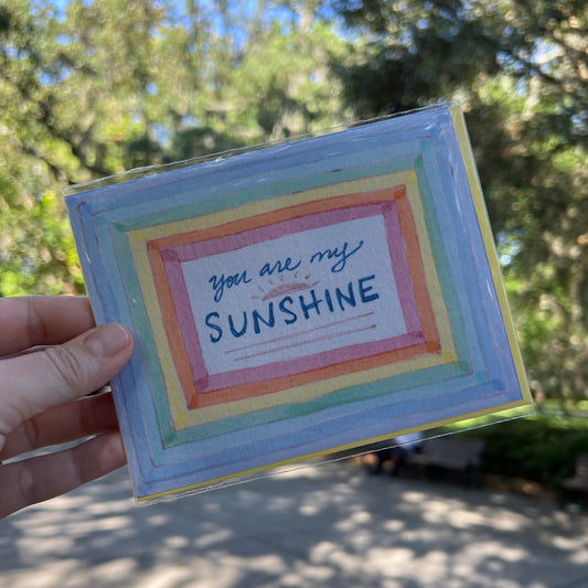 A hand holding a square, hand-painted card or art print with a rainbow border and the words 'you are my SUNSHINE' written on it, set against a blurred outdoor background of trees and sunlight.