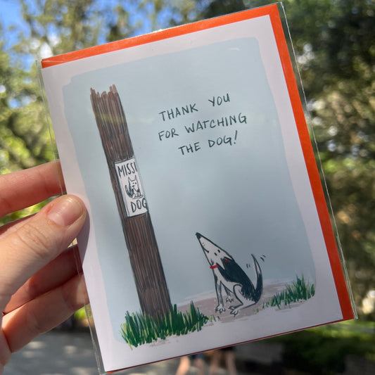 A hand holding a greeting card outdoors. The card features a humorous illustration of a dog on a leash next to a 'MAY NOT DOG' warning sign, with the text 'THANK YOU FOR WATCHING THE DOG!' on a light blue background with an orange border.