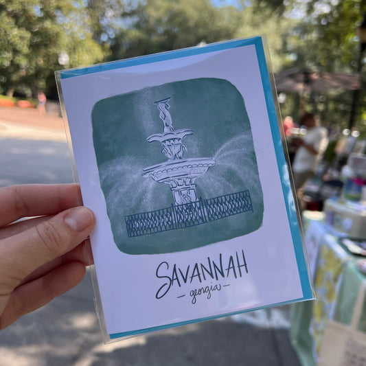 A hand holding a printed card or postcard outdoors. The card has a white background with a blue border, a green painted square containing a fountain illustration, and the word 'SAVANAH' printed on the right side.