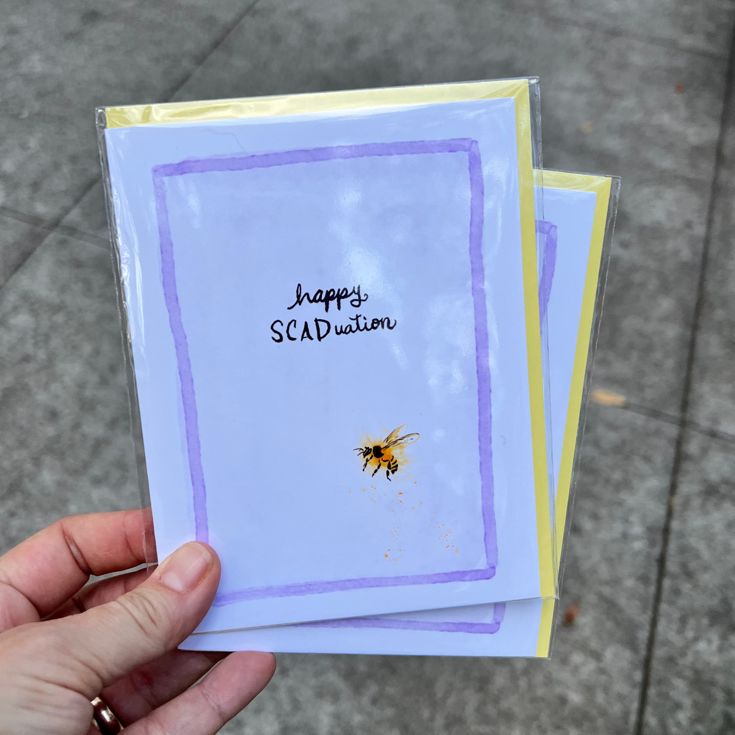 Hand holding a stack of cards with a bee illustration and 'happy SCADuation' text on a pavement background.
