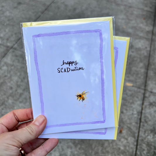 Hand holding a stack of cards with a bee illustration and 'happy SCADuation' text on a pavement background.