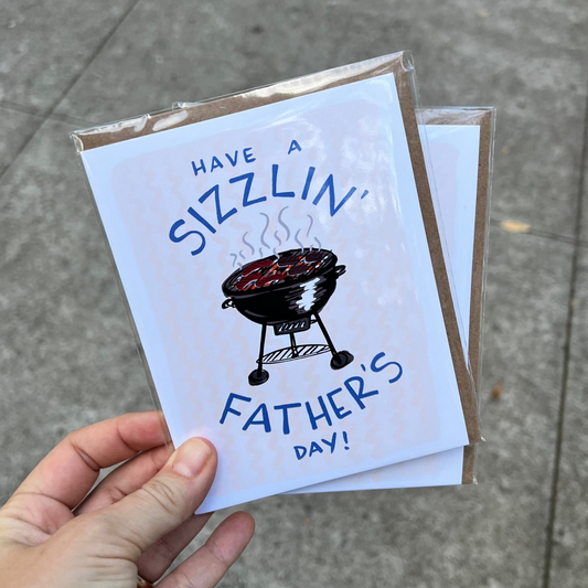 Hand holding a Father's Day card with a grill illustration on a concrete surface