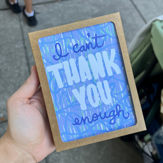 A hand holds a square framed art print with the text 'I can't THANK YOU enough' in white and blue lettering on a textured blue background, set in a natural wood frame.